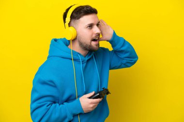 Sarı arka planda izole edilmiş bir bilgisayar oyunu kontrolörüyle oynayan beyaz bir adam bir şeyler dinlerken elini kulağına koyar.