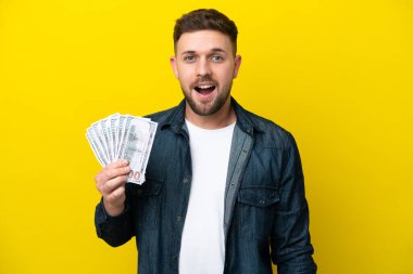 Kafkasyalı genç bir adam, sarı arka planda, şaşırtıcı bir yüz ifadesiyle izole edilmiş bir sürü para alıyor.