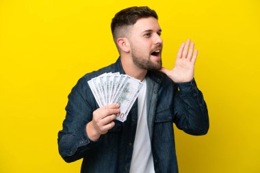 Kafkasyalı genç adam, sarı arka planda izole edilmiş, ağzı açık bir şekilde bağırarak bir sürü para alıyor.