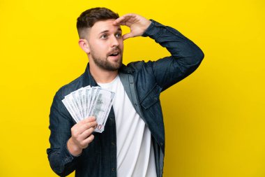 Kafkasyalı genç bir adam sarı arka planda izole edilmiş bir sürü para alıyor. Yan tarafa bakarken sürpriz jest yapıyor.