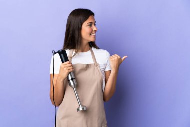 Mor arka planda izole edilmiş el blenderi kullanan Brezilyalı kadın bir ürünü sunmak için yan tarafı işaret ediyor