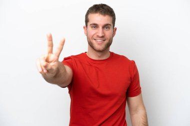 Beyaz arka planda izole edilmiş genç beyaz adam gülümsüyor ve zafer işareti gösteriyor.