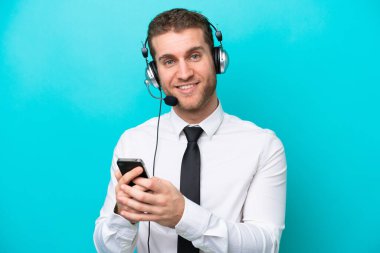 Tele pazarlamacı beyaz adam mavi arka planda izole edilmiş bir kulaklıkla çalışıyor cep telefonuyla bir mesaj gönderiyor.