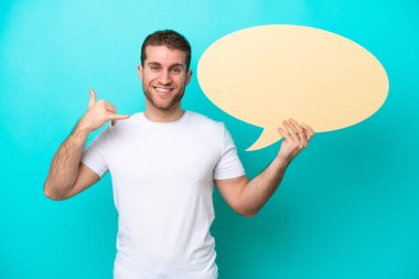 Kafkasyalı genç adam mavi arka planda izole edilmiş boş bir konuşma balonu tutuyor ve telefon jesti yapıyor.