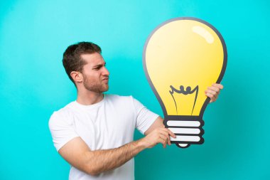Mavi arka planda izole edilmiş genç beyaz bir adam elinde bir ampul simgesi tutuyor ve kuşkuları var.