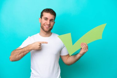 Kafkasyalı genç adam mavi arka planda izole edilmiş bir şekilde elinde bir kontrol simgesi tutuyor ve ona doğrultuyor.