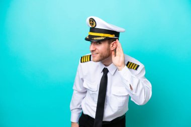 Kafkasyalı uçak pilotu mavi arka planda izole edilmiş bir şekilde elini kulağına koyarak bir şey dinliyor.