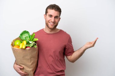 Beyaz arka planda izole edilmiş alışveriş torbası tutan genç beyaz bir adam davet etmek için elini yana uzatıyor.