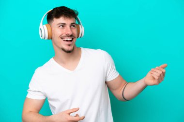 Beyaz tenli yakışıklı adam mavi arka planda izole edilmiş müzik dinliyor ve gitar hareketi yapıyor.