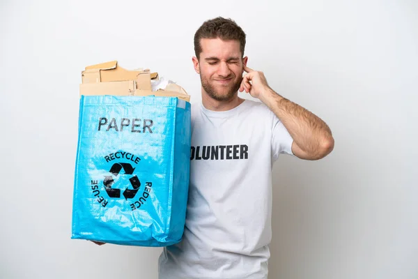 年轻的高加索人拿着一个装满纸的回收袋 沮丧地从白色背景中分离出来 捂住耳朵进行回收 — 图库照片