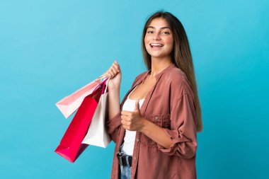 Mavi arka planda izole edilmiş genç beyaz kadın elinde alışveriş torbaları ve başparmağıyla