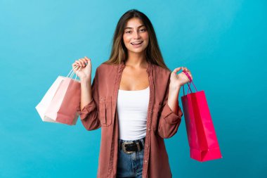 Mavi arka planda izole edilmiş genç beyaz bir kadın alışveriş torbaları tutuyor ve gülümsüyor.