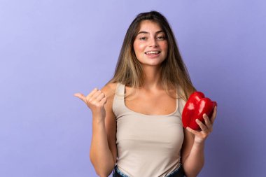 Mor arka planda izole edilmiş biber tutan genç bir kadın bir ürünü sunmak için kenarı işaret ediyor.