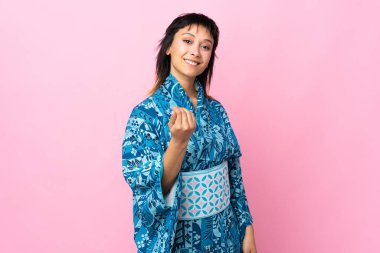 İzole edilmiş mavi arka planda kimono giyen genç bir kadın benimle gelmesi için davet ediyor. Gelmene sevindim.