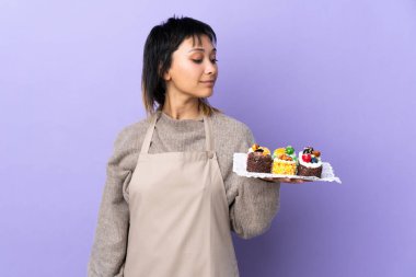 Genç Uruguaylı kadın bir sürü farklı mini keki izole edilmiş mor arka planda tutuyor.