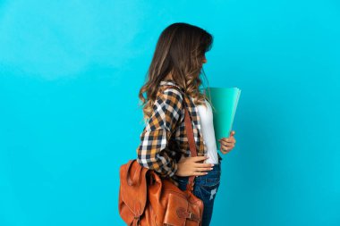 Genç bir bayan öğrenci, mavi arka planda izole edilmiş ve çaba gösterdiği için sırt ağrısı çekiyor.