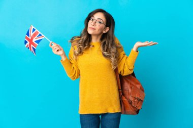 Birleşik Krallık bayrağını tutan genç İspanyol kadın mavi arka planda izole edilmiş bir şekilde ellerini kaldırırken şüpheleri var.