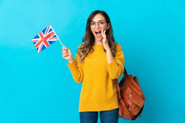 Birleşik Krallık bayrağını tutan genç İspanyol kadın mavi arka planda izole edilmiş ağzı açık bağırıyor.