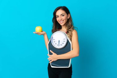 Mavi arka planda tartı tutan beyaz bir kadın. Tartı makinesiyle ve elmayla izole edilmiş.
