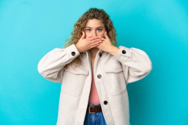 Mavi arka planda izole edilmiş genç sarışın kadın elleriyle ağzını kapatıyor.