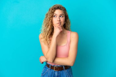 Mavi arka planda izole edilmiş genç sarışın kadın yukarı bakarken bir fikir düşünüyor.