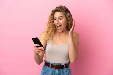 Pembe arka planda izole edilmiş genç sarışın kadın kameraya bakarken şaşırmış bir ifadeyle cep telefonunu kullanıyor.