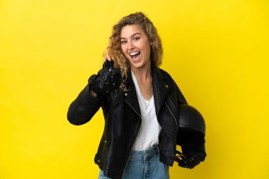 Motosiklet kasklı genç sarışın kadın sarı arka planda izole edildi. Başparmakları havada çünkü iyi bir şey oldu.