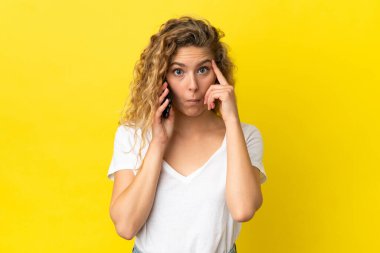 Sarı arka planda izole edilmiş cep telefonu kullanan genç sarışın bir kadın bir fikir düşünüyor.