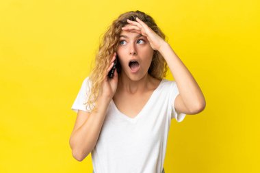 Sarı arka planda izole edilmiş cep telefonu kullanan genç sarışın kadın yan tarafa bakarken sürpriz jest yapıyor.