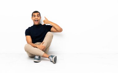 Afro-Amerikalı bir adam yerde oturuyor ve telefon jesti yapıyor. Bana geri dönüş işareti ver.