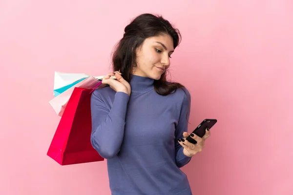 十代のロシアの女の子はピンクの背景に隔離されたショッピングバッグを保持し 友人に彼女の携帯電話でメッセージを書く — ストック写真