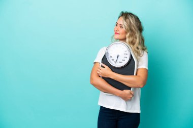 Genç Brezilyalı bir kadın mavi arka planda izole edilmiş, tartı makinesi ve diğer tarafa bakıyor.