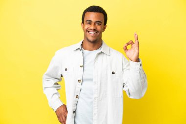 İzole edilmiş sarı arka planda Afro-Amerikalı yakışıklı adam parmaklarıyla tamam işareti gösteriyor.
