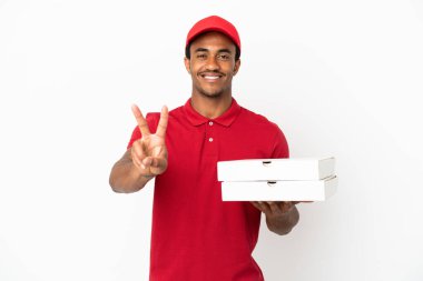 Afro-Amerikalı pizza teslimatçısı izole edilmiş beyaz duvardan pizza kutuları alıyor gülümsüyor ve zafer işareti gösteriyor.