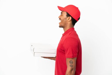 Afro-Amerikalı pizzacı, izole edilmiş beyaz duvardan pizza kutularını alıyor ve yan pozisyonda gülüyor.