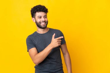 Faslı genç bir adam sarı arka planda izole edilmiş. Parmakla göstererek bir ürün sunuyor.