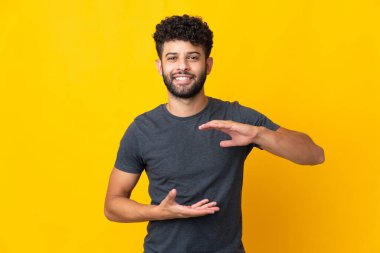 Faslı genç bir adam sarı arka planda izole edilmiş. Elinde bir ilan vermek için avucunda hayali bir alan tutuyor.