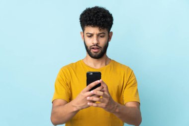 Genç Faslı adam mavi arka planda izole edilmiş kameraya bakarken şaşırmış bir ifadeyle cep telefonuna bakıyor.