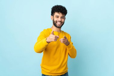 Faslı genç bir adam mavi arka planda izole edilmiş şaşırmış ve ön tarafı işaret ediyor.