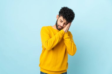 Faslı genç bir adam mavi arka planda izole edilmiş bir şekilde uyku jesti yapıyor.