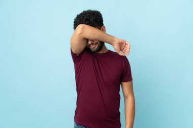 Faslı genç bir adam mavi arka planda izole edilmiş. Gözlerini elleriyle kapatıyor.