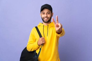 Morarmış arka planda izole edilmiş spor çantalı genç Faslı adam gülümsüyor ve zafer işareti gösteriyor.