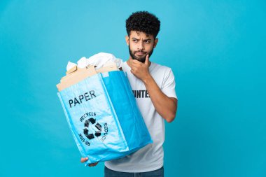 Genç Faslı bir adam, izole edilmiş arka plan üzerinde geri dönüşüm yapmak için kağıt dolu bir geri dönüşüm çantası tutuyor. Şüpheleri ve düşünceleri var.