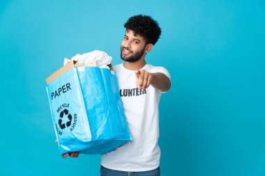 Genç Faslı bir adam, izole edilmiş arkaplanı geri dönüştürmek için elinde kağıt dolu bir geri dönüşüm çantası tutuyor ve mutlu bir ifadeyle ön tarafı işaret ediyor.