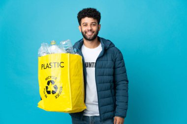 Elinde plastik şişelerle dolu bir çanta tutan genç Faslı adam izole edilmiş arka planı geri dönüştürmek için kolları kalçasında poz veriyor ve gülümsüyor.