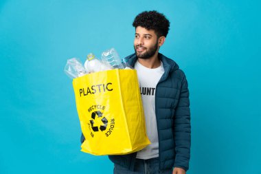 Genç Faslı bir adam, izole edilmiş arka planda geri dönüşüm için plastik şişelerle dolu bir çanta tutuyor. Yan tarafa bakıyor ve gülümsüyor.