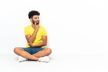 Genç Faslı yakışıklı adam izole edilmiş arka planda yerde oturuyor. Yukarı bakarken bir fikir düşünüyor.