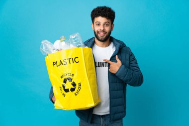 Genç Faslı bir adam, izole edilmiş arkaplanı süpriz bir yüz ifadesiyle geri dönüştürmek için plastik şişelerle dolu bir çanta tutuyor.