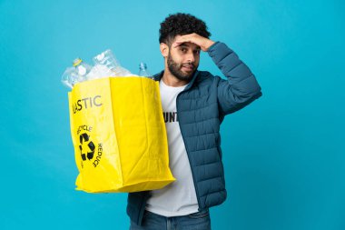 Genç Faslı bir adam, izole edilmiş arkaplanı geri dönüştürmek için plastik şişelerle dolu bir çanta tutuyor. Yan tarafa bakarken sürpriz jest yapıyor.