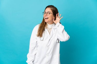 Orta yaşlı Brezilyalı doktor, mavi arka planda izole edilmiş bir şekilde elini kulağına koyarak bir şey dinliyor.